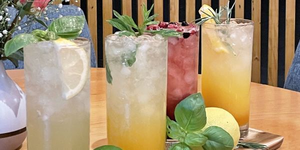 Vier bunte Cocktails auf einem Holztablett, dekoriert mit frischen Kräutern und Früchten.