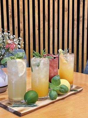 Vier bunte Cocktails auf einem Holztablett, dekoriert mit frischen Kräutern und Früchten.