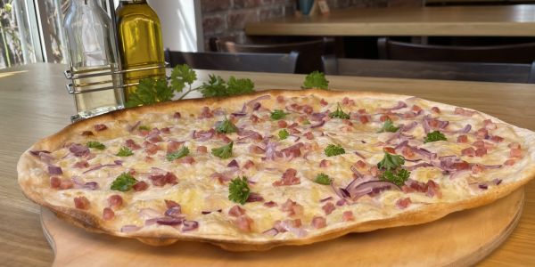 Flammkuchen mit Speck und Zwiebeln auf Holzbrett, dekoriert mit Petersilie und Ölflaschen.